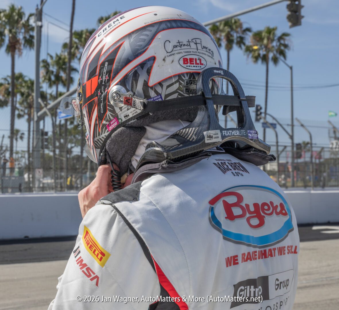 A race car driver in full racing gear adjusts their helmet near a track, with palm trees and safety fencing in the background.