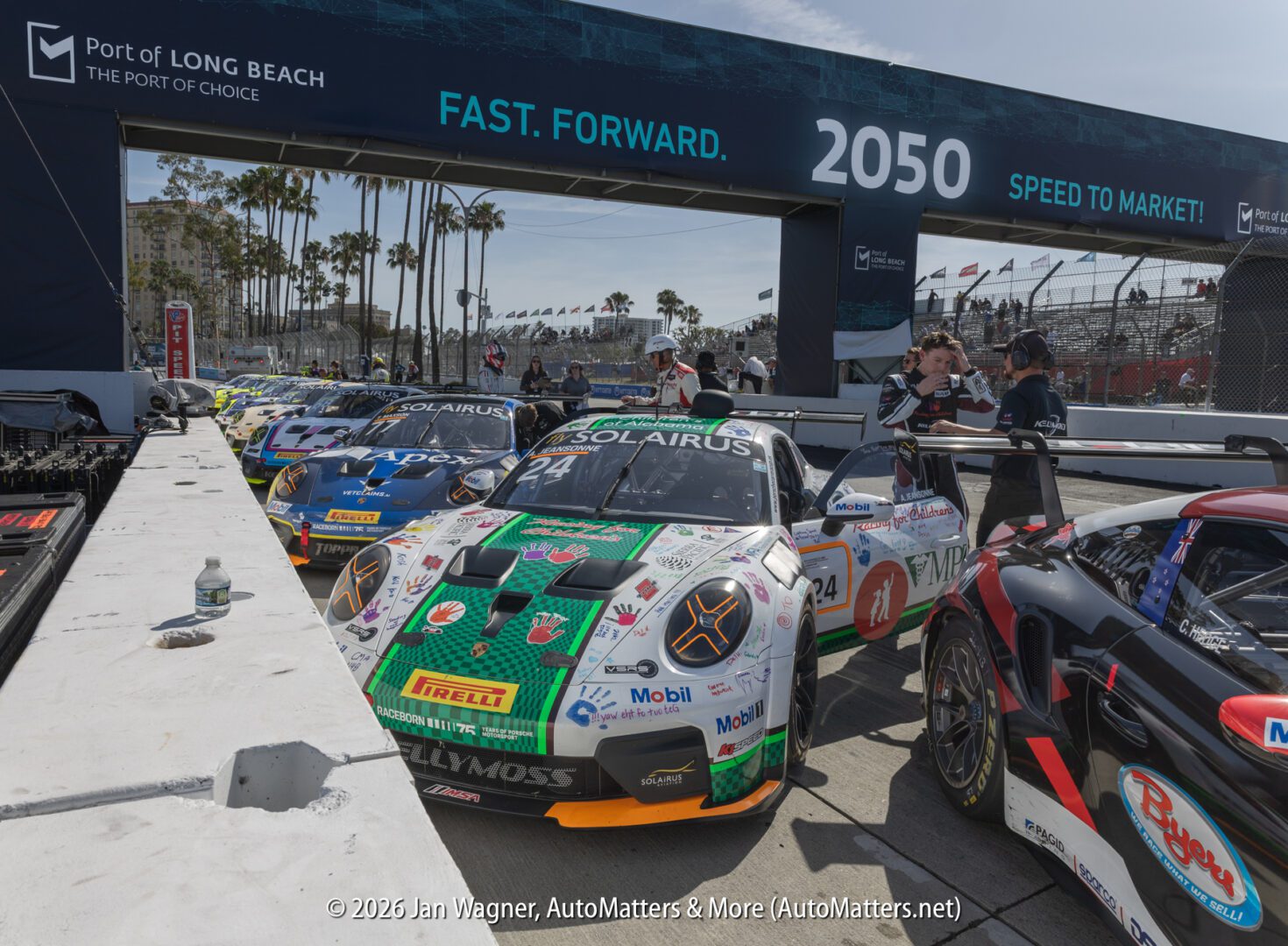 Several race cars lined up on a track at the starting grid of the Long Beach Grand Prix, with banners and palm trees visible in the background.