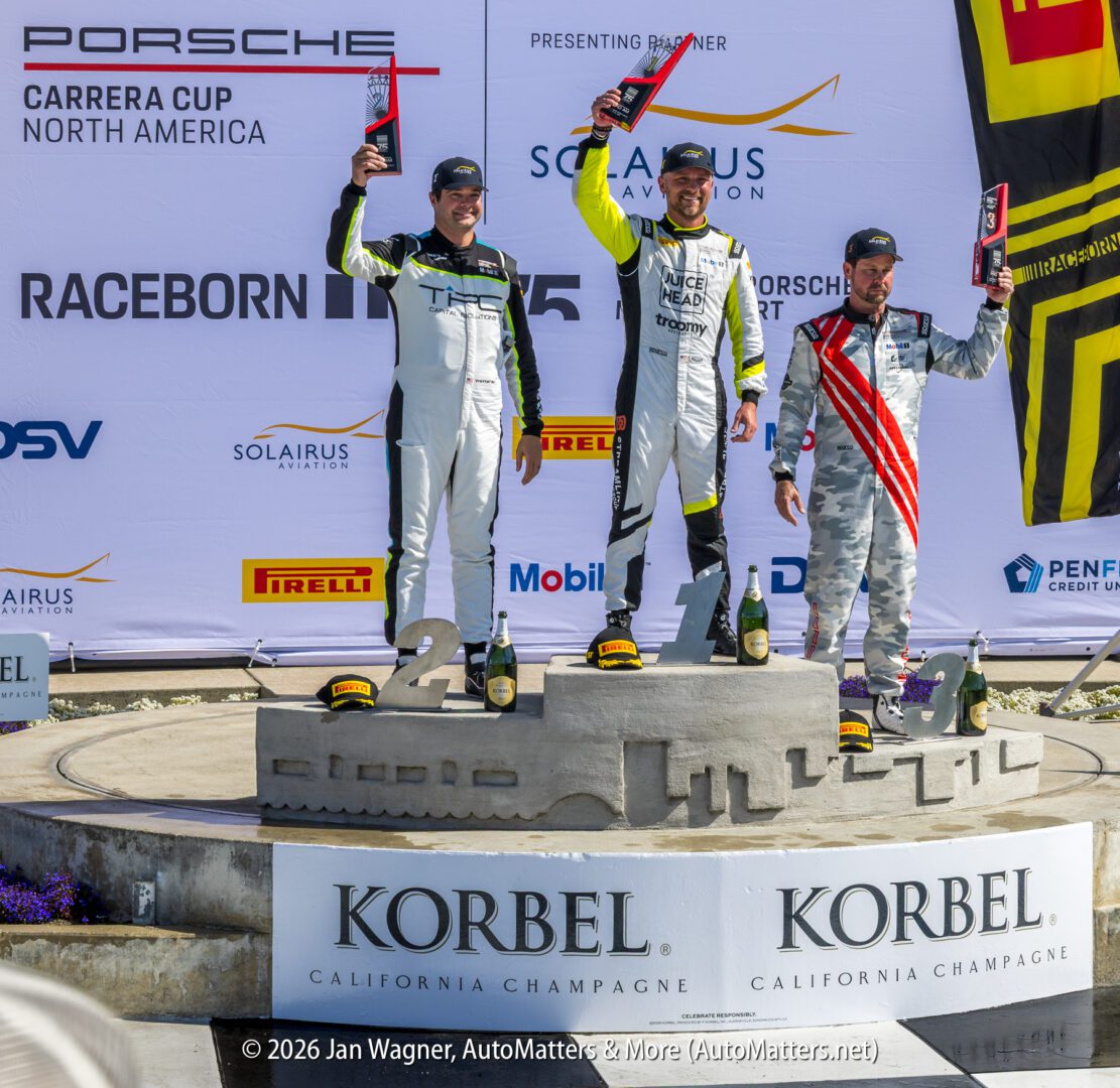 Three race car drivers in racing suits stand on a podium holding trophies, celebrating their win at a Porsche Carrera Cup North America event.