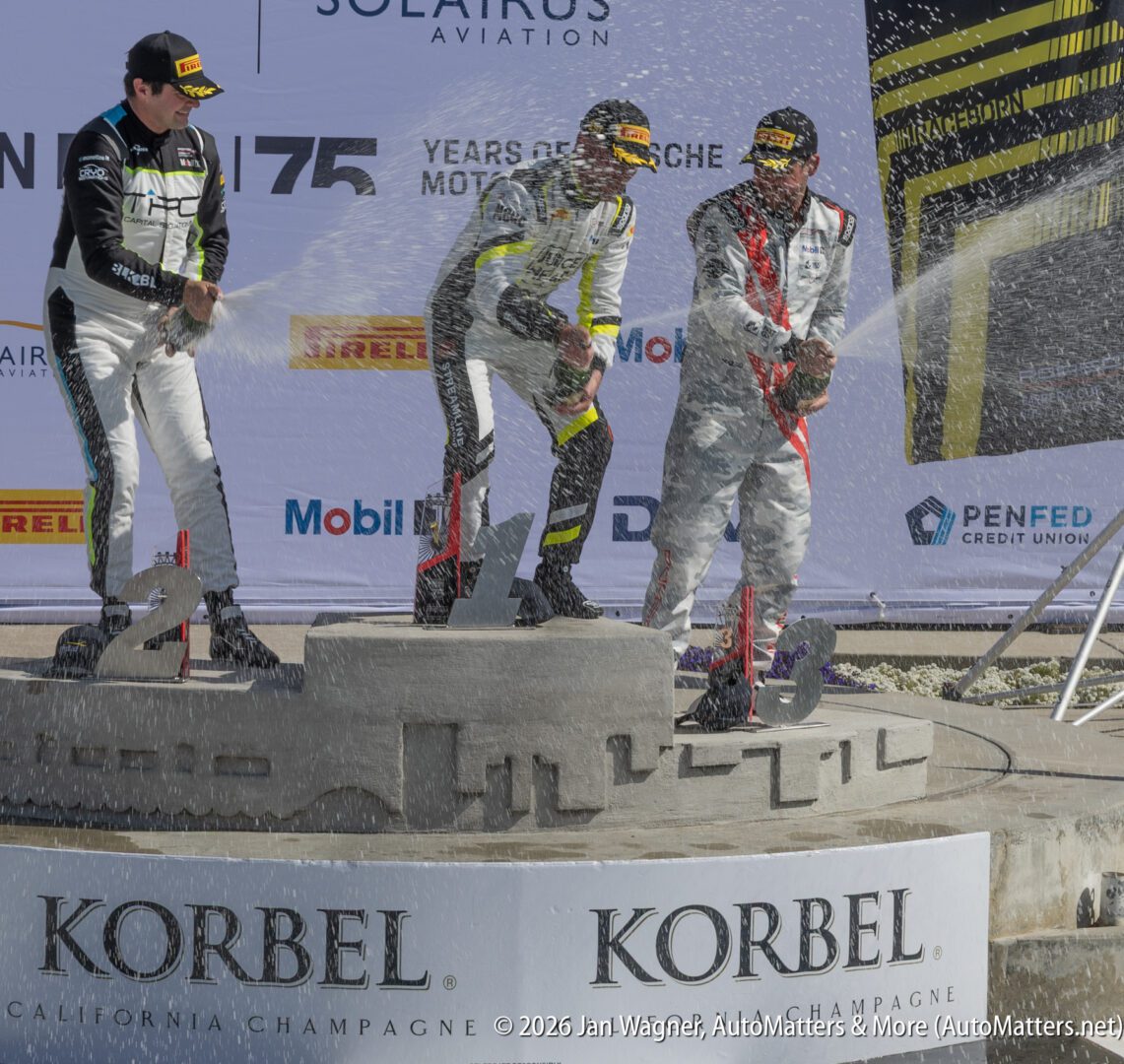 Three race car drivers spray champagne on a podium during a motorsport awards ceremony, with branded banners and a sandcastle-shaped stage.
