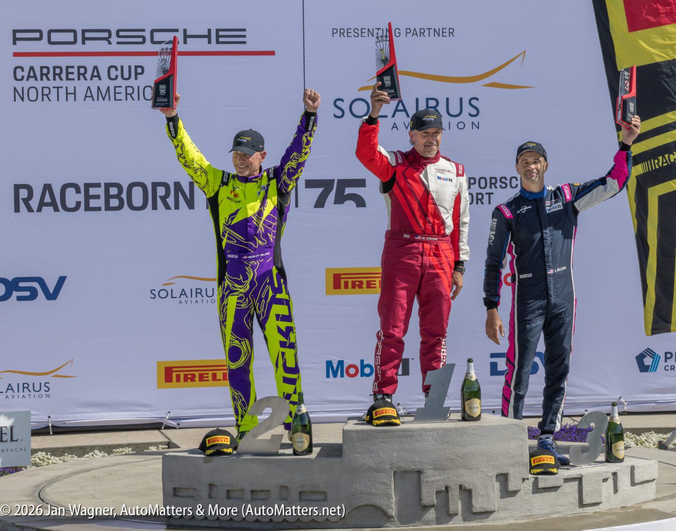 Three race car drivers in racing suits stand on a podium holding trophies, celebrating their victory at a Porsche Carrera Cup North America event.