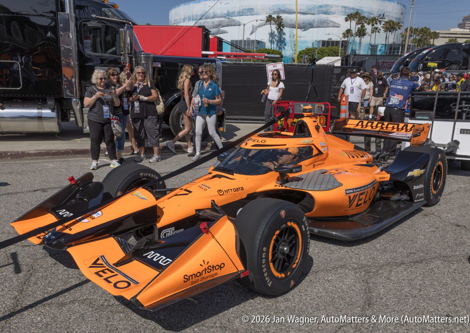 An orange IndyCar with VELO branding is parked as fans and photographers gather around, with palm trees and transport trucks in the background.