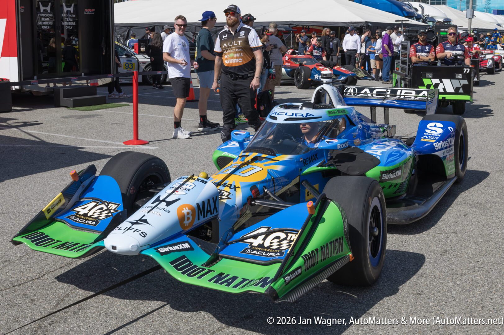 A blue, green, and white Acura IndyCar sits on a racetrack paddock with several people standing nearby and other race cars in the background.