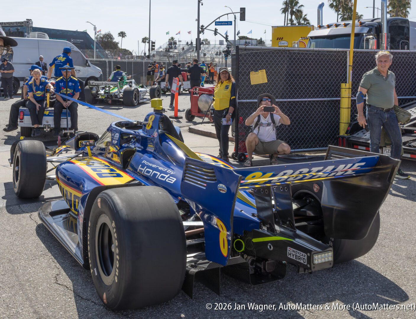 A blue IndyCar with number 27 is parked as people stand nearby and take photos in a paddock area at a motorsports event.