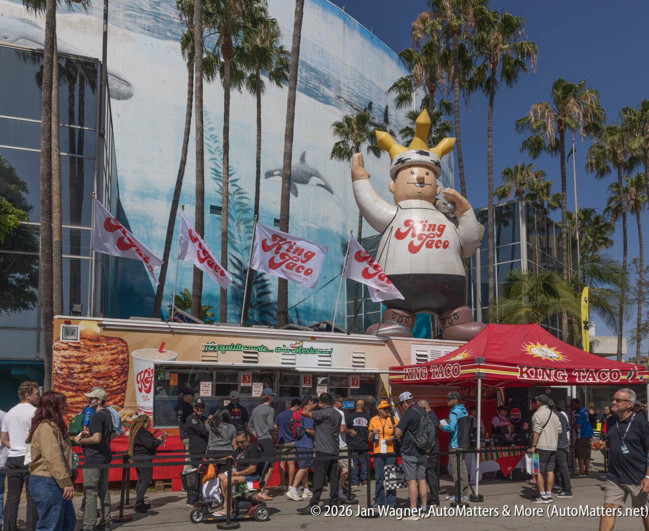 People line up at a King Taco food truck with a large inflatable mascot outside an aquarium mural featuring dolphins and surrounded by palm trees.