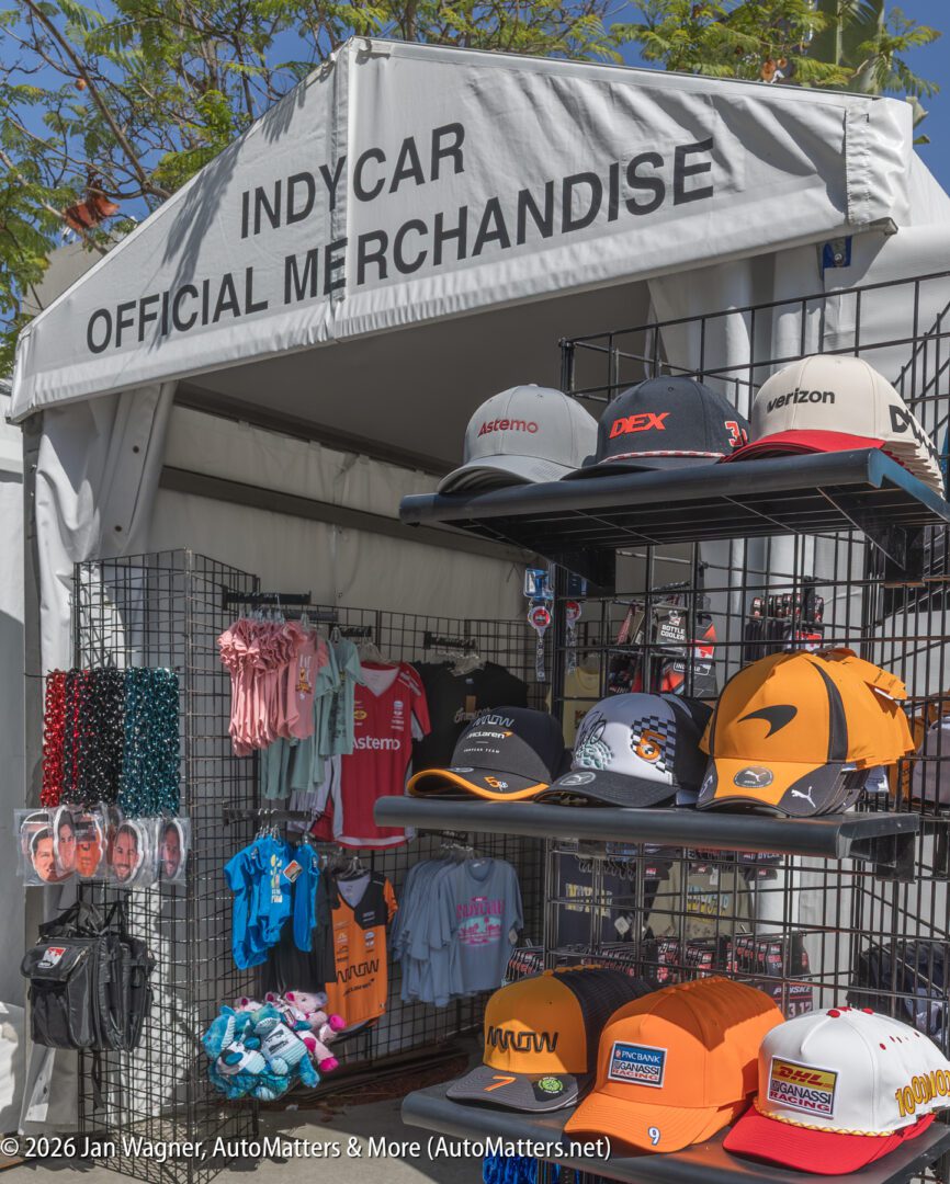 Outdoor display of IndyCar official merchandise, including hats, shirts, lanyards, and keychains, set up in front of a branded white canopy tent.