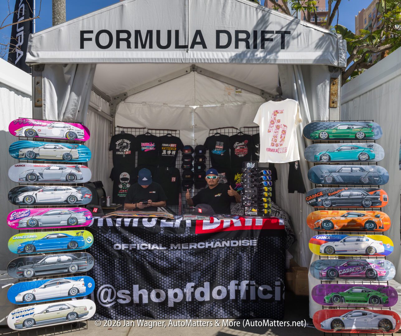 A Formula Drift merchandise booth displays t-shirts, hats, and colorful skateboard decks with car designs, with two people seated behind the counter.