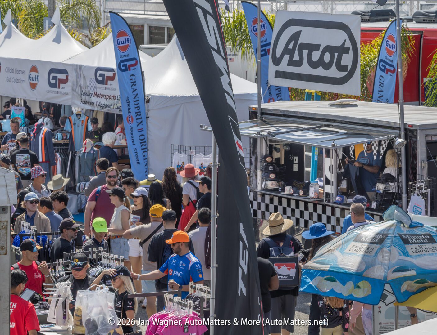 A crowd browses merchandise booths and vendor displays at an outdoor motorsports event, with branded flags and signs visible overhead.