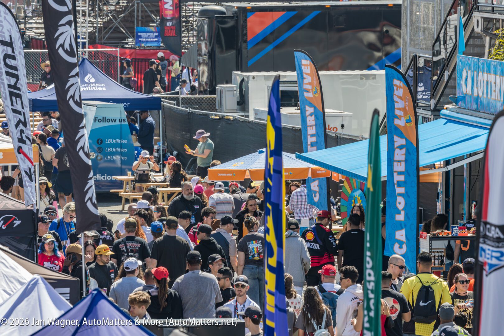 A large crowd of people walks through an outdoor event area lined with vendor tents, flags, and banners on a sunny day.