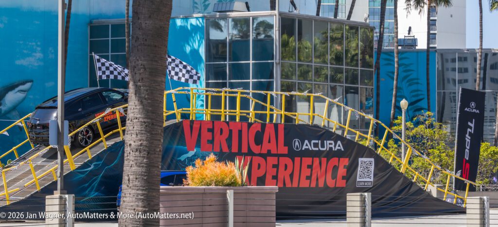 A black Acura SUV drives up a steep yellow ramp at an outdoor Acura Vertical Xperience event, with checkered flags and promotional banners in view.