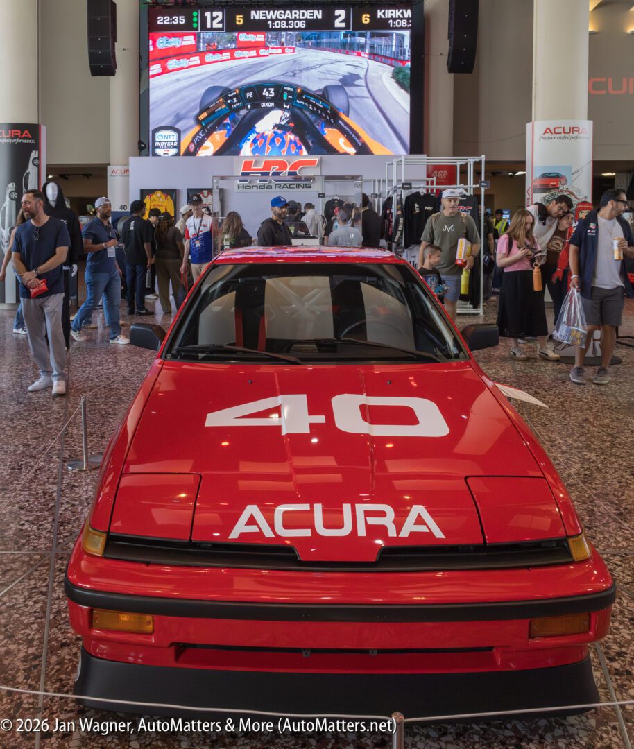 A red Acura car with the number 40 displayed on the hood is showcased indoors; people and racing merchandise are in the background, with a racing event shown on a large screen above.