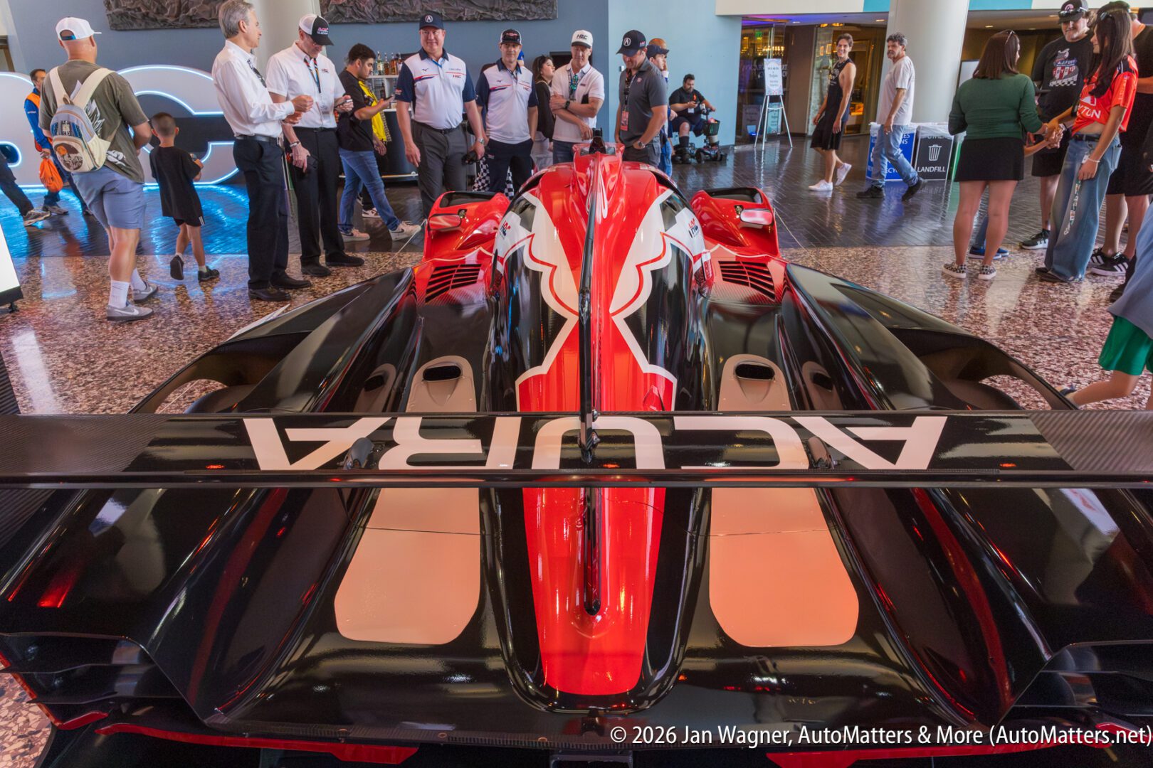 A red and black Acura race car on display indoors, surrounded by people taking photos and observing, under bright lighting.
