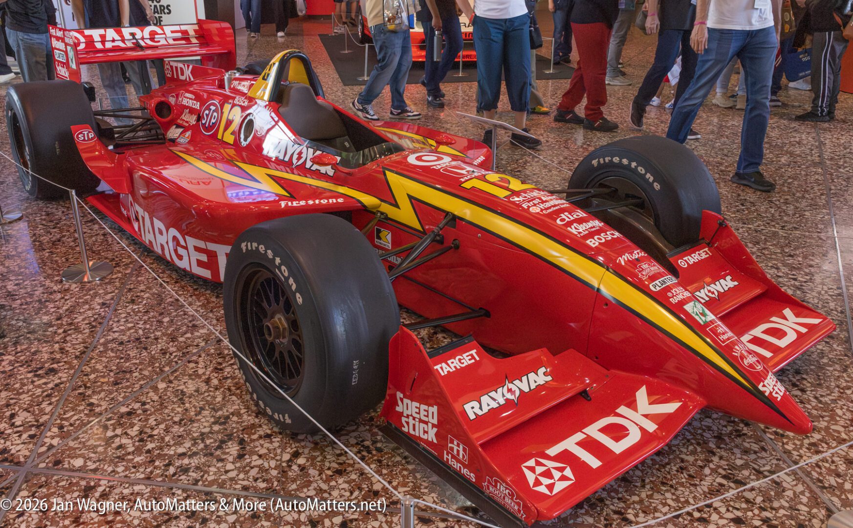 A red Target-branded IndyCar race car with yellow lightning accents is displayed indoors on a polished floor, surrounded by people.