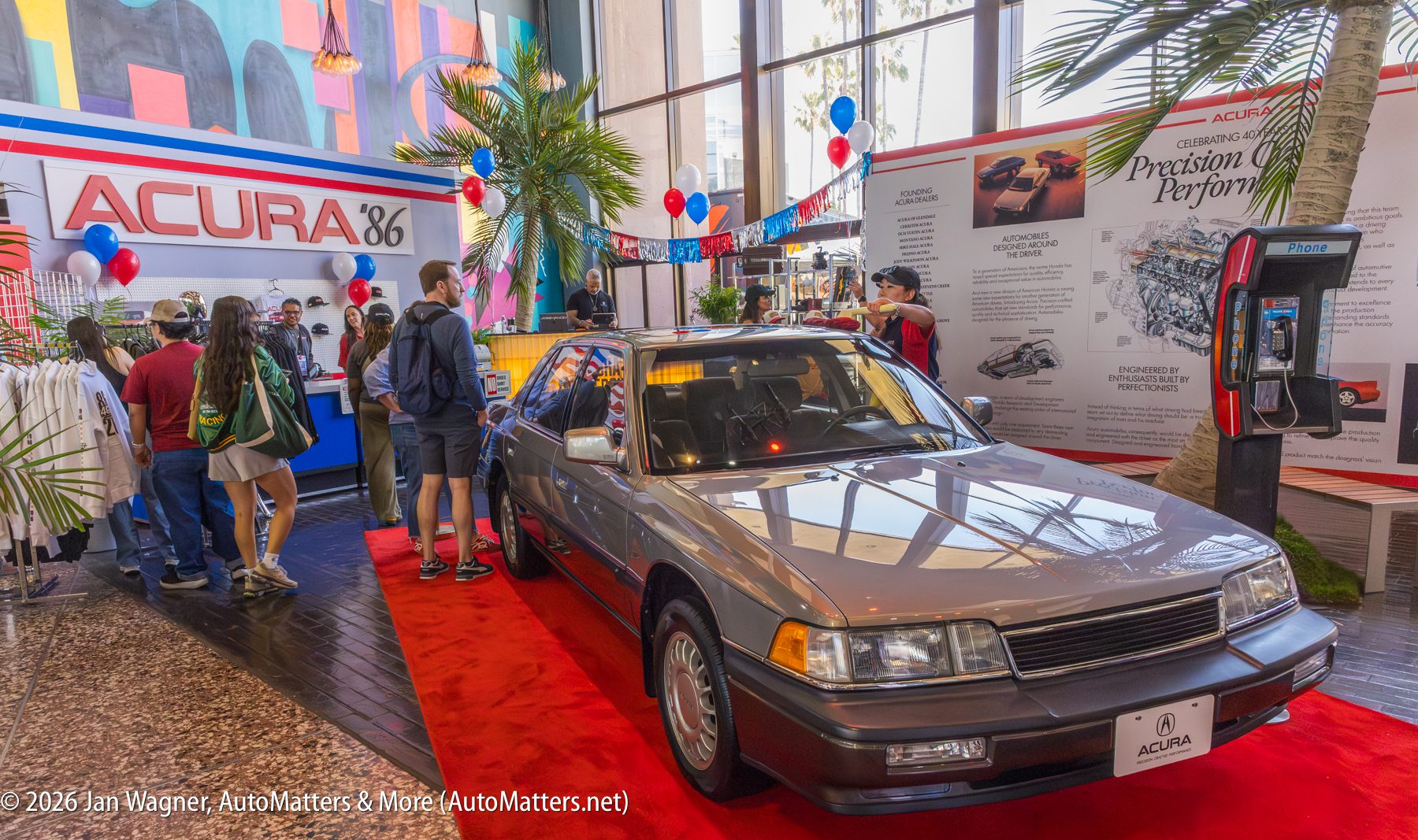 A classic Acura sedan is displayed indoors at an Acura promotional event, with people browsing nearby exhibits and merchandise.