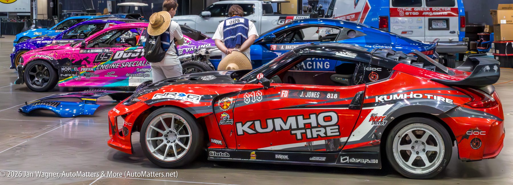 Three modified race cars with colorful decals are parked indoors; two people in hats are inspecting the vehicles.