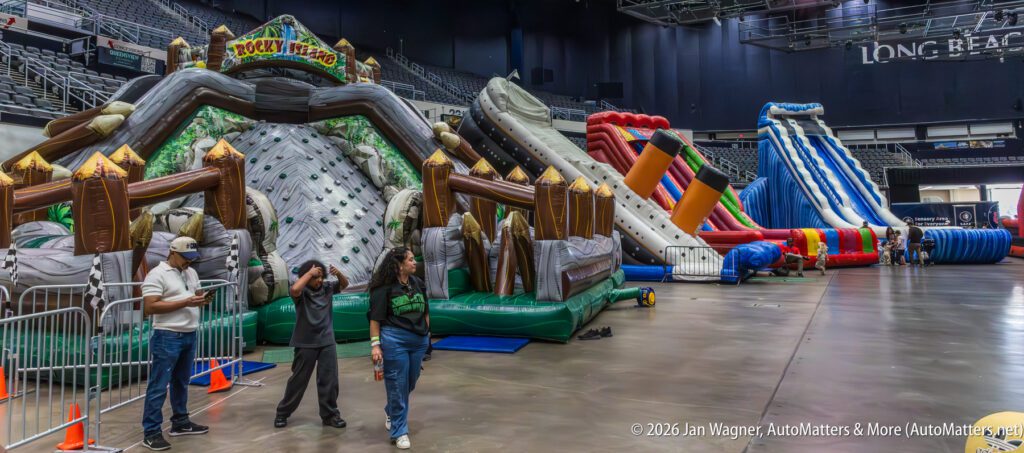Several large inflatable slides and obstacle courses are set up inside an indoor arena, with a few people standing and walking nearby.