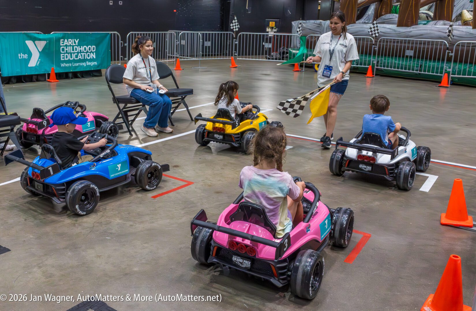 Children in small electric cars line up at a starting line indoors, with an adult holding a checkered flag and another adult seated nearby. Orange cones mark the course.