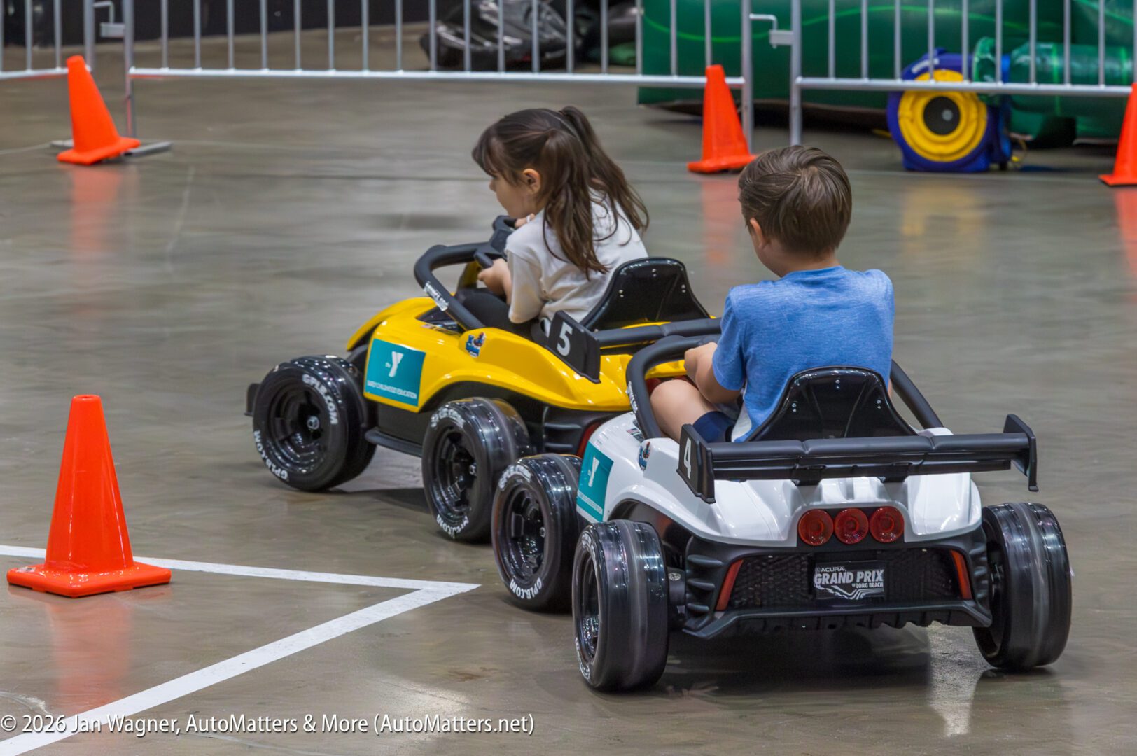 Two children drive small electric cars around an indoor track marked with orange cones.