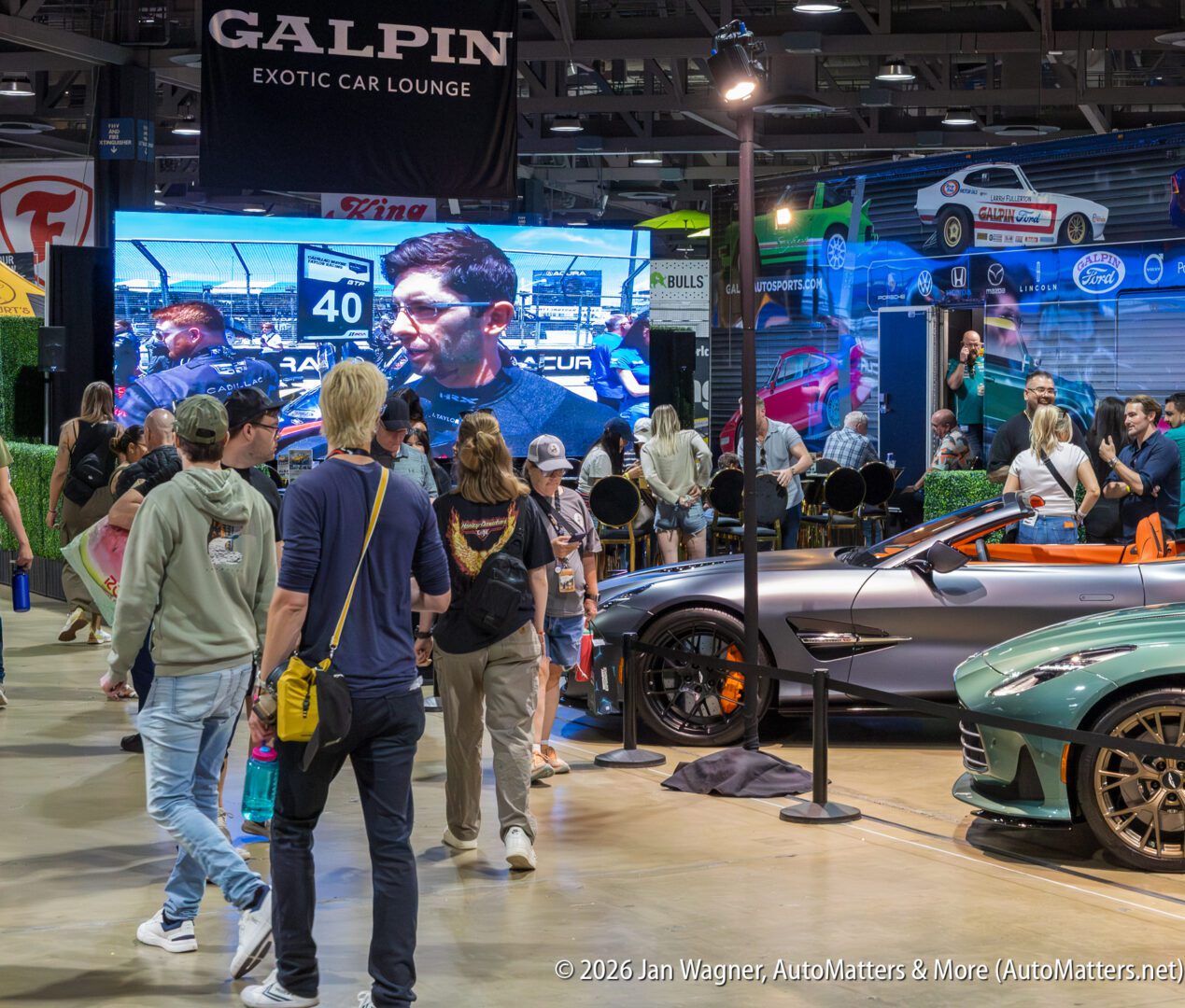 People walking through an indoor car show with luxury cars on display and a large screen showing a racing video in the background.