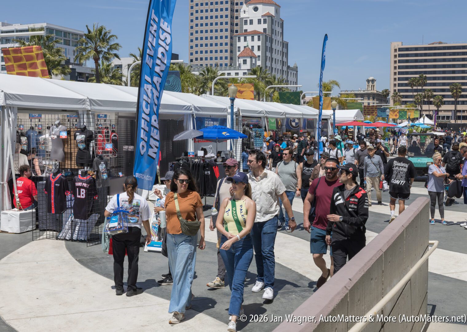 Crowd of people walk past merchandise booths at an outdoor event with city buildings and palm trees in the background. Official merchandise signs are visible.