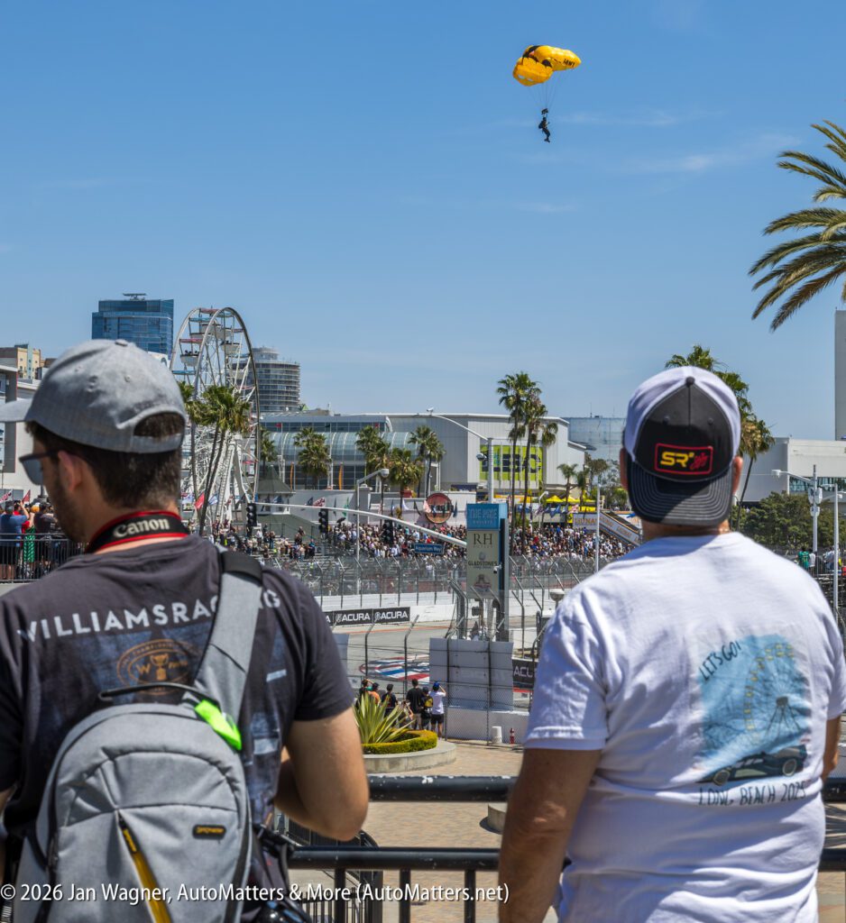 Two spectators watch a car race on a city track while a parachutist descends from the sky. A Ferris wheel and palm trees are visible in the background.
