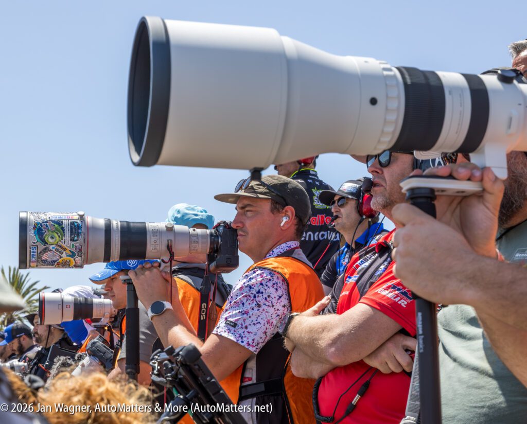 A group of photographers with large telephoto lenses aim their cameras at an outdoor event under a clear sky.