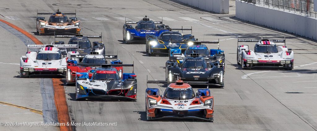 A group of race cars compete on a paved track during a sports car race, with several cars closely packed as they navigate a turn.