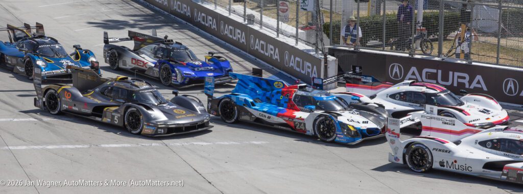 Several sports cars are lined up on a race track, preparing to start, with Acura branding visible along the barriers.