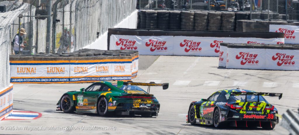 Two race cars navigate a turn on a city street circuit, passing barriers and sponsor signs, with tire stacks lining the track edge.