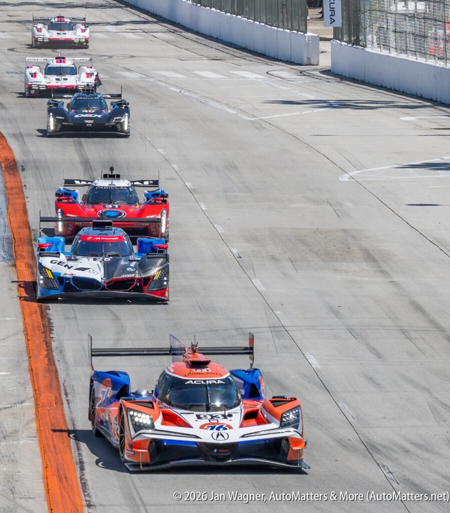 Six prototype race cars drive in a line on a paved racetrack during a daytime motorsport event, with barriers and fencing visible alongside the track.