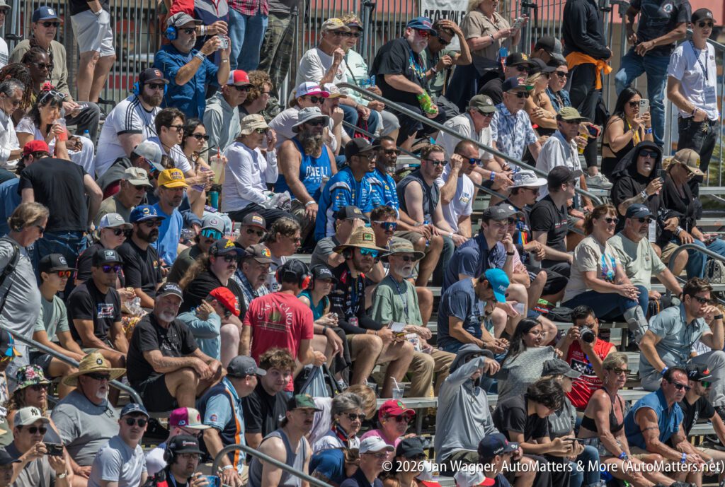 A large crowd of people sits in outdoor stadium bleachers on a sunny day, many wearing hats and sunglasses.