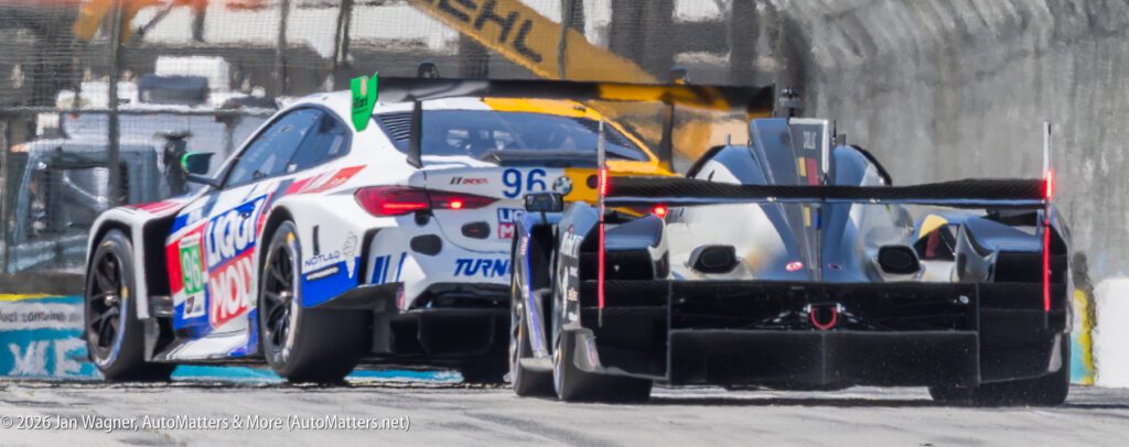 A white BMW M4 GT3 and a black prototype race car drive side by side on a racetrack, with blurred fencing and barriers in the background.