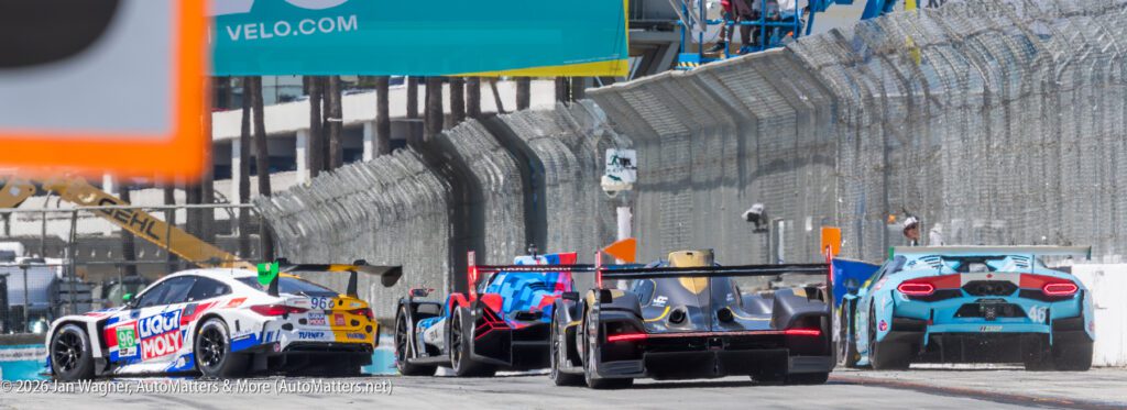 Four race cars drive on a fenced street circuit, turning right under an overhead VELO.com sign during a motorsport event.