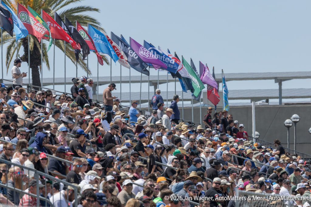 A large crowd sits in bleachers under colorful sponsor flags at an outdoor event on a sunny day.