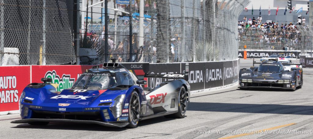 Two Cadillac race cars drive on a city street circuit during a professional motorsport event, with barriers and advertisements lining the track.