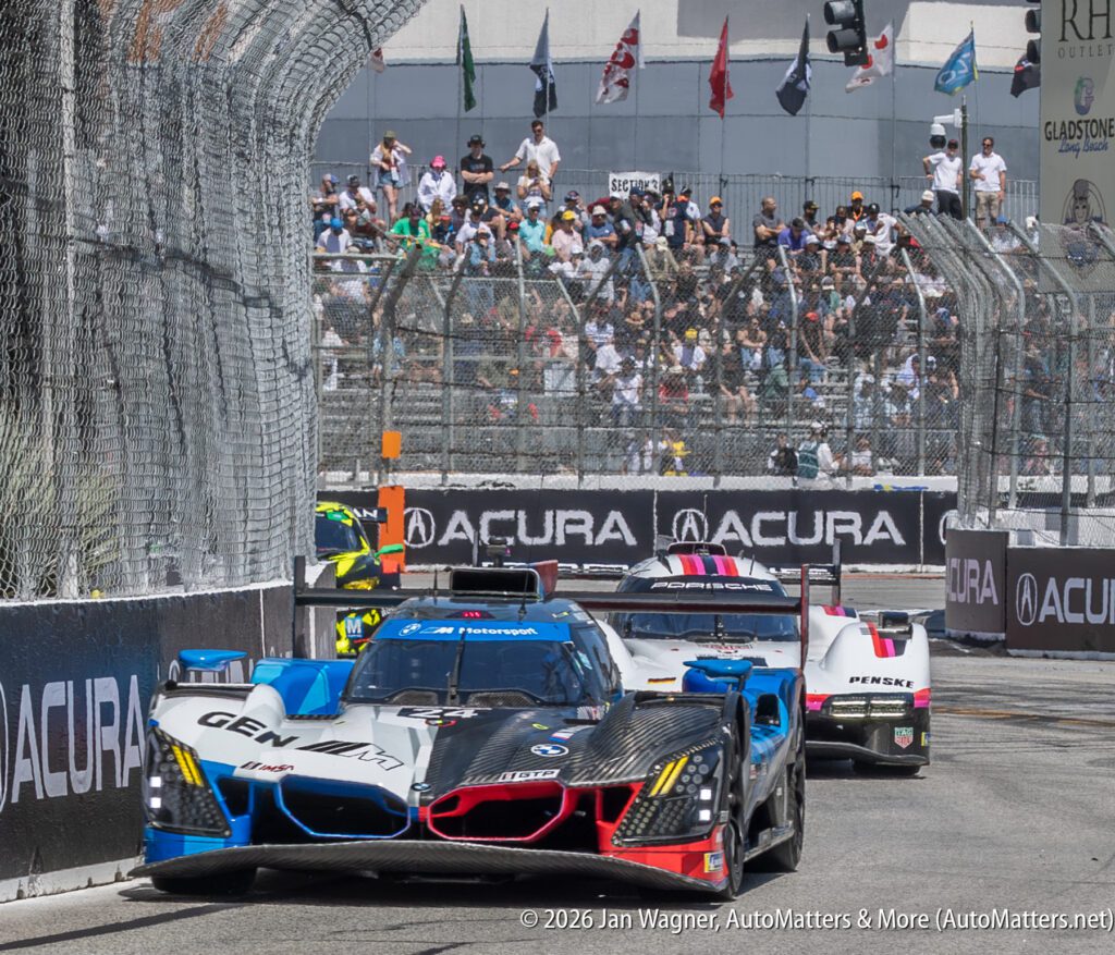Three race cars compete on a city street circuit during an Acura-sponsored motorsport event, with barriers and spectators in the background.