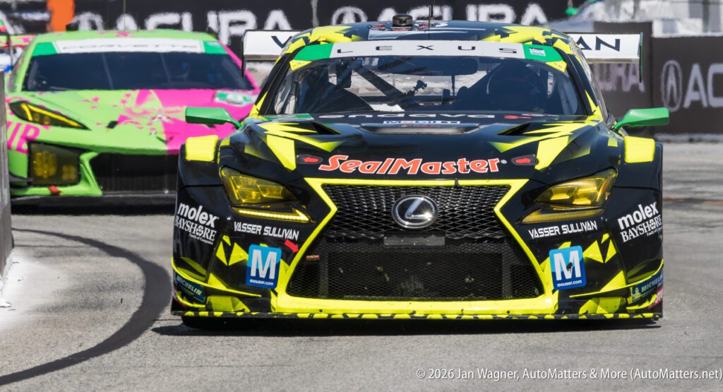 A black and yellow Lexus race car with SealMaster branding leads on a racetrack, with a pink and green car following behind.