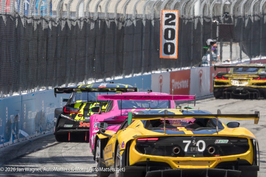 Four race cars drive close together on a fenced street circuit, approaching a sign marked "200" during a motorsport event.
