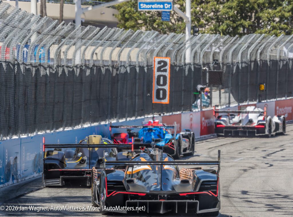 Several prototype race cars drive closely together on a fenced street circuit, approaching a sign marked "200" under the "Shoreline Dr" street sign.
