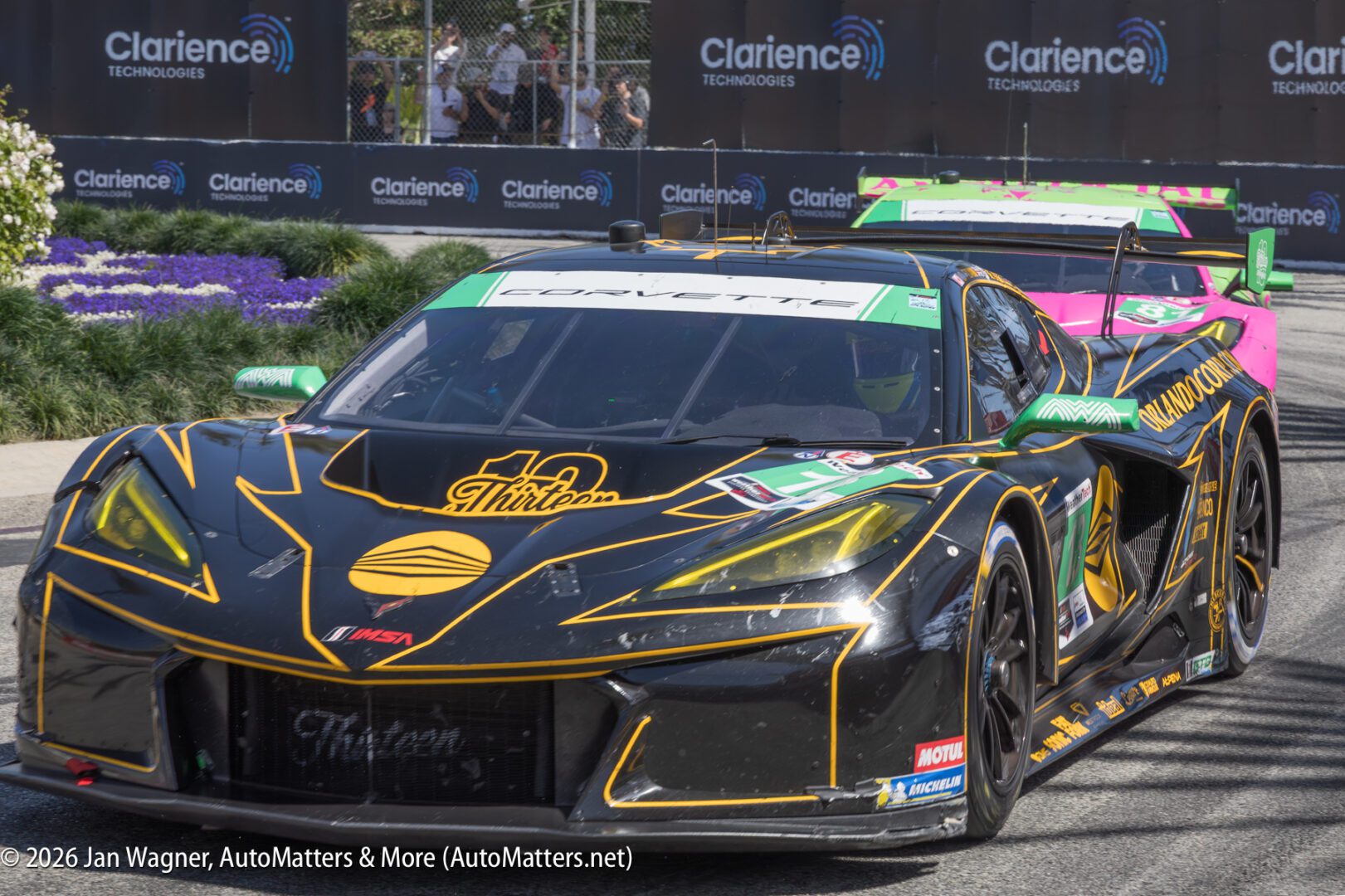 A black and yellow Corvette race car leads a bright pink car on a city street circuit, with spectators and sponsor banners in the background.