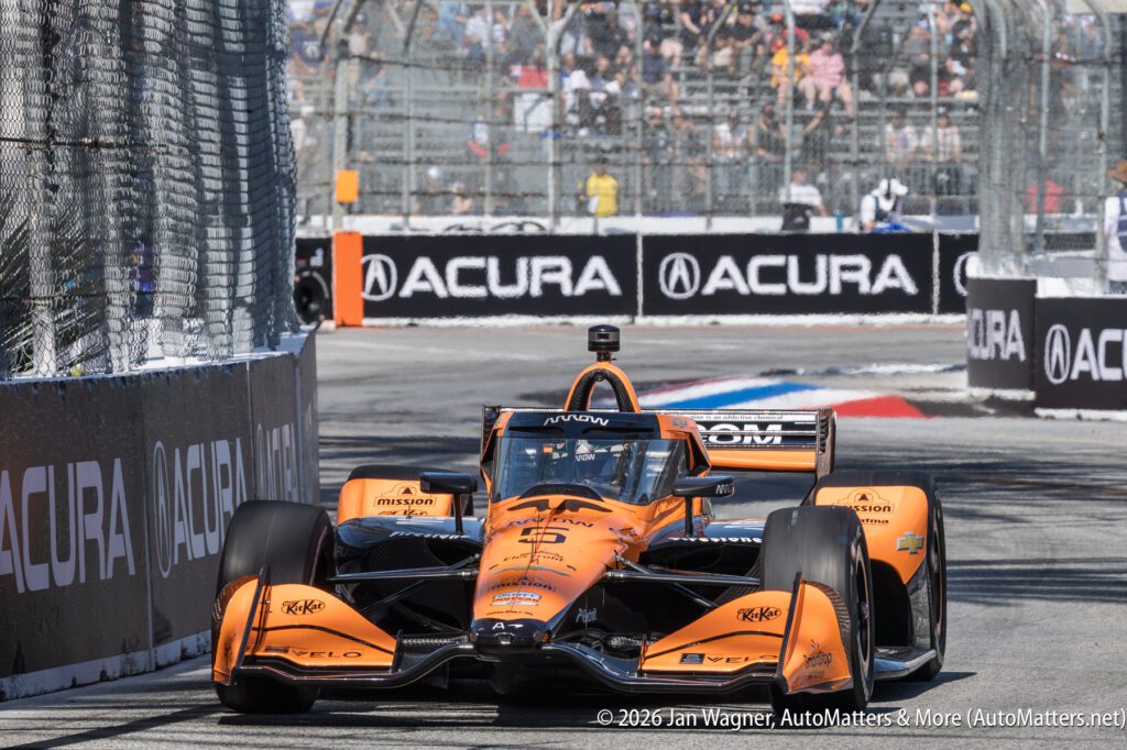 An orange IndyCar races on a street circuit, passing Acura-branded barriers, with spectators watching from behind a fence in the background.