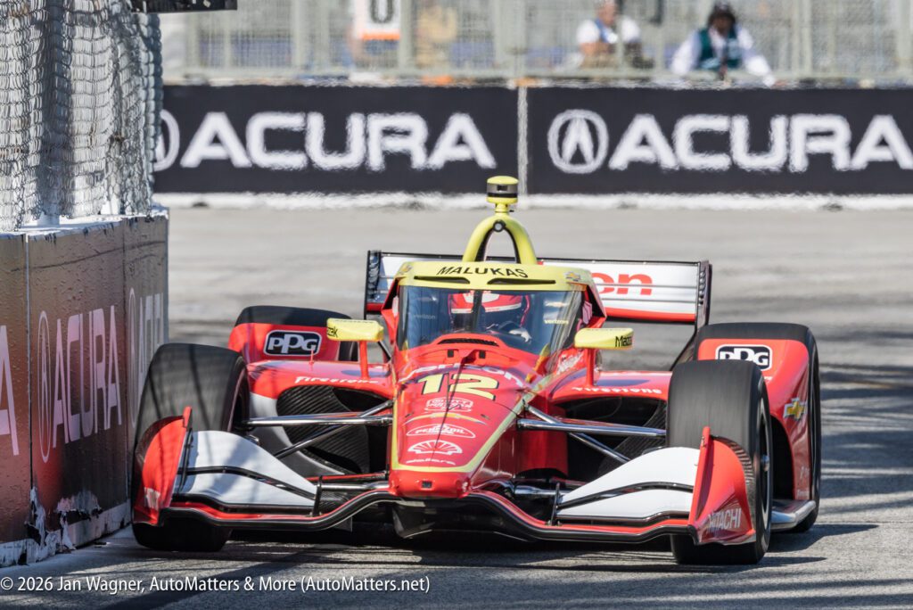 A red IndyCar with number 18, labeled "Malukas," drives on a city racetrack near an Acura-branded barrier during a race.