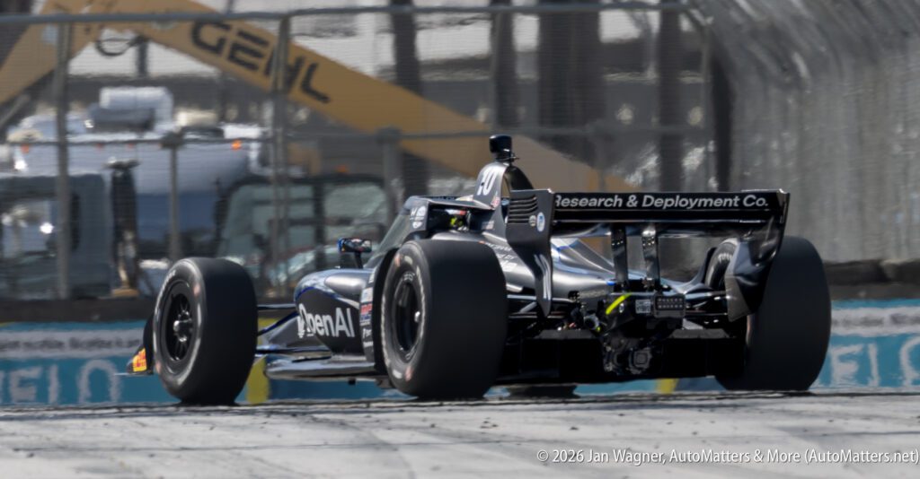 A race car labeled "openAI" drives on a city racetrack, with construction equipment and safety fencing visible in the background.