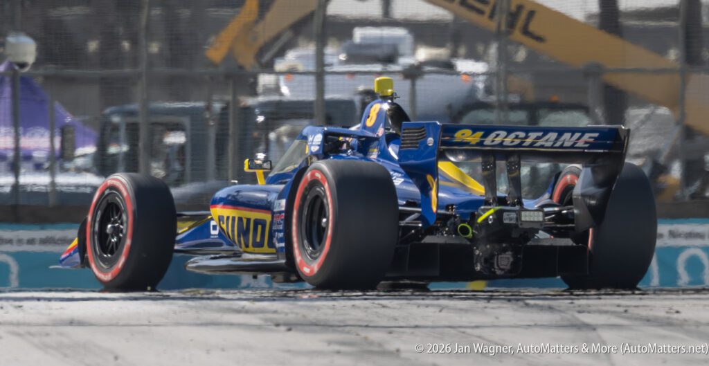A blue and yellow IndyCar races on a track, kicking up debris, with construction equipment visible in the background.
