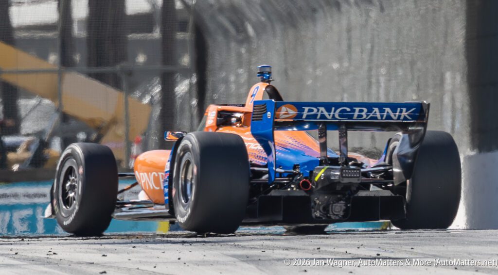 A rear view of an orange and blue PNC Bank-sponsored IndyCar driving on a racetrack, with debris and tire marks visible on the asphalt.