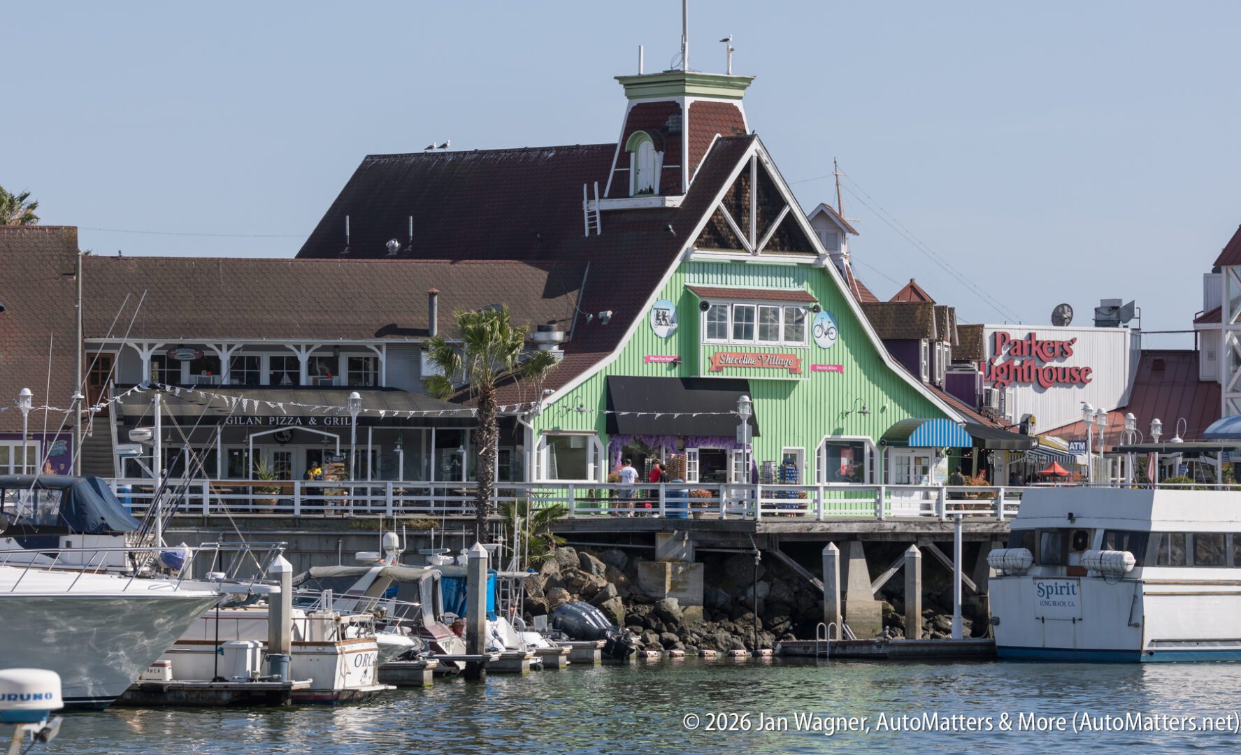 A waterfront marina scene with boats docked in front of colorful buildings and restaurants, including a green and white structure and Parker’s Lighthouse.