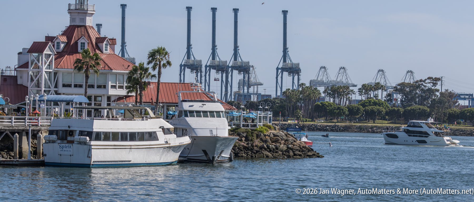 Several boats are docked near a red-roofed building with palm trees, with large industrial cranes visible in the background across the water.