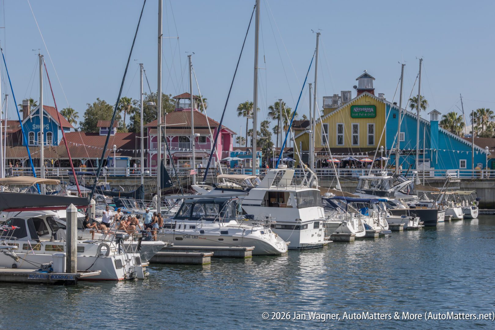 Several boats and yachts are docked in a marina with colorful buildings and palm trees in the background under a clear sky.