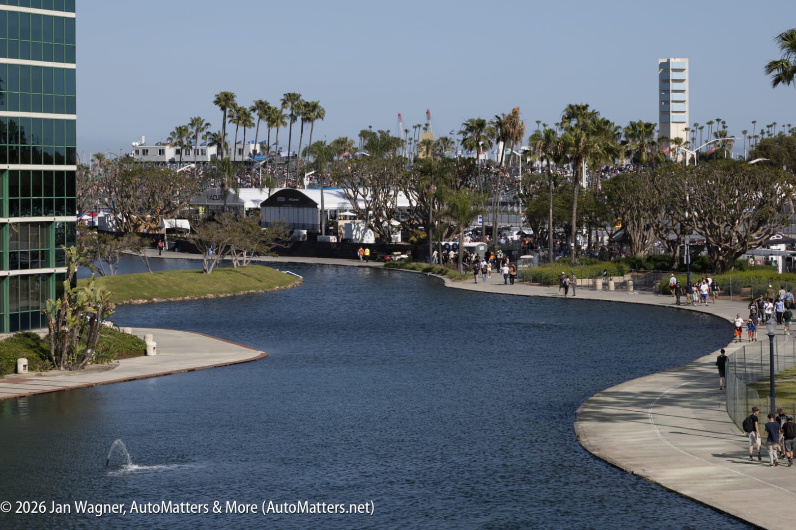 A view of a curved waterfront walkway lined with palm trees, with people walking and gathering in the distance near modern buildings.