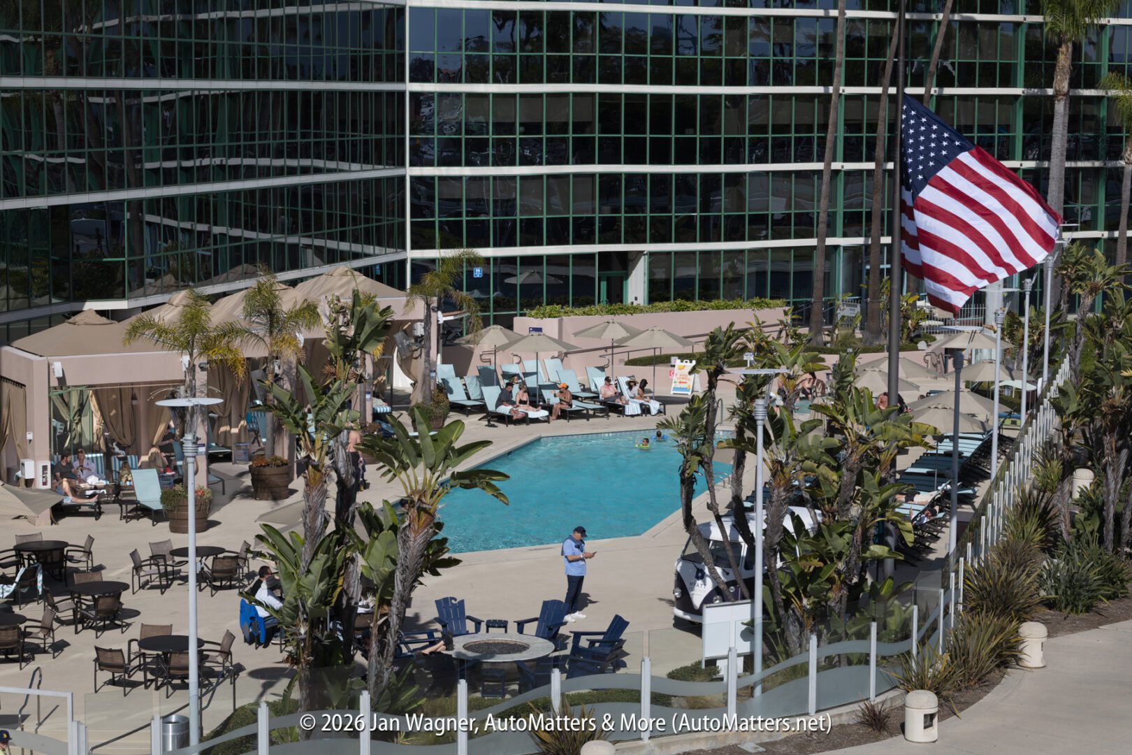An outdoor hotel pool area with lounge chairs, cabanas, palm trees, and a large American flag, surrounded by a modern glass building.